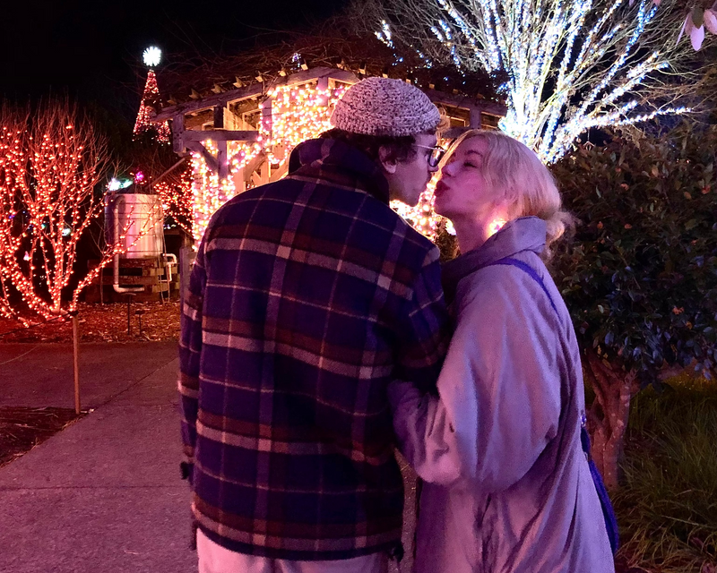 Two people embracing in a festive outdoor setting with lights and trees.