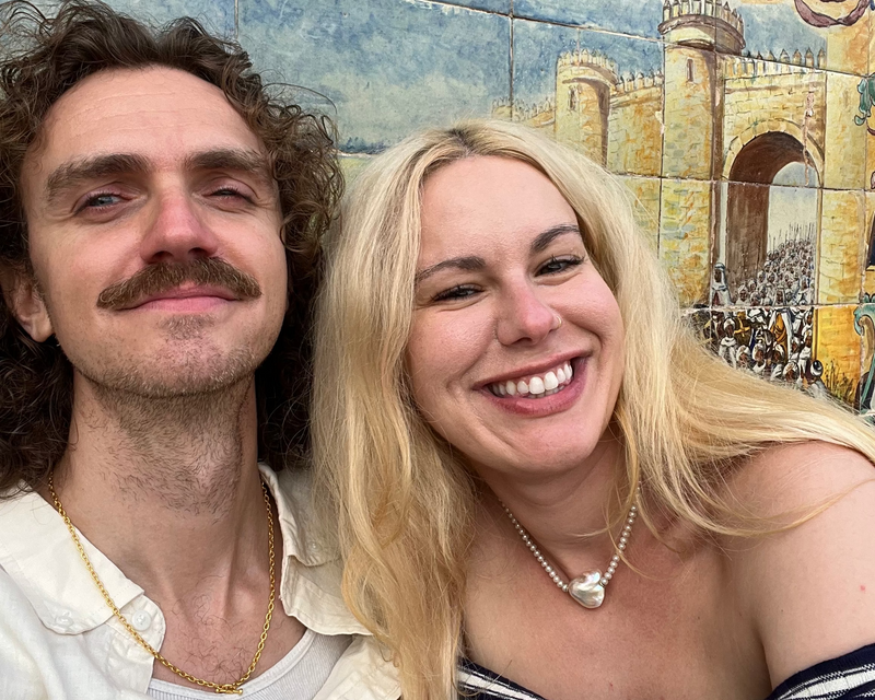 Two people posing in front of a colorful mural with architectural elements.