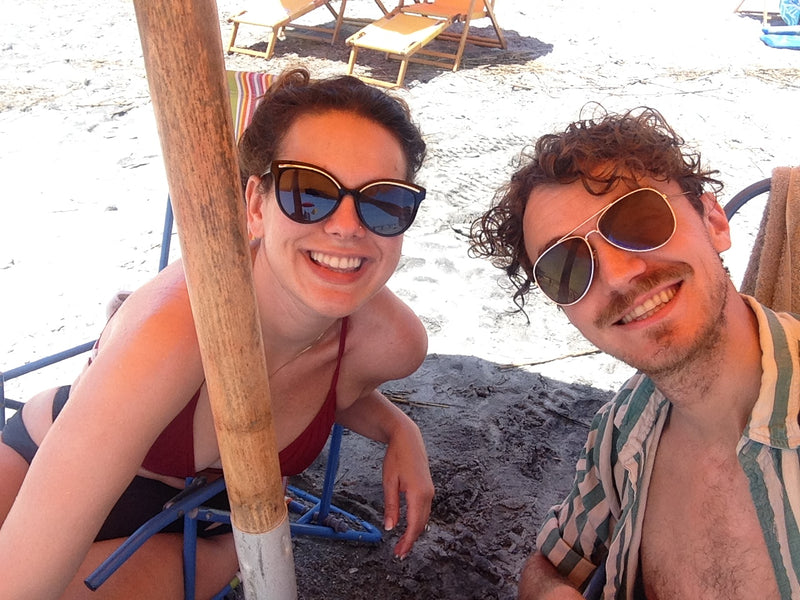 Two people on a beach with sunglasses, sitting under a straw umbrella.