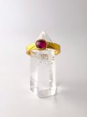 A 24K gold-banded ring with a large red rubystone on top, displayed on a clear crystal stand.