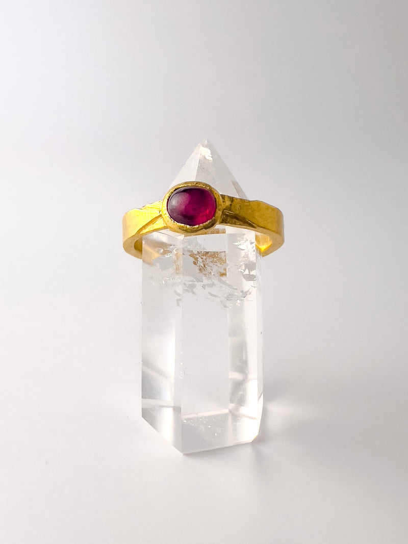 A 24K gold-banded ring with a large red rubystone on top, displayed on a clear crystal stand.